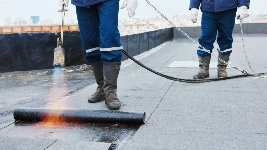 Roofers torch-applying modified bitumen waterproofing membrane on a flat roof